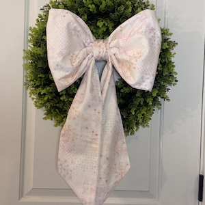 May include: A decorative wreath with a dense green base and a large, patterned bow. The bow features a soft pink and white floral design. The wreath is hanging on a white door, creating a welcoming aesthetic. The bow's fabric has a delicate pattern.