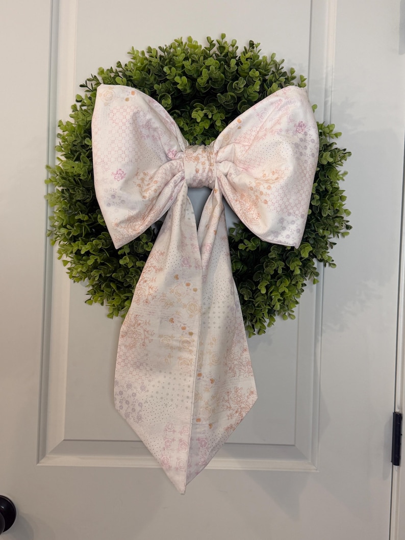 May include: A decorative wreath with a lush green base and a large, patterned bow. The bow features a soft pink and white floral design, adding a touch of elegance. The wreath is hanging on a white door, creating a welcoming aesthetic.