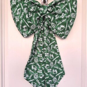 May include: A large green bow with a repeating pattern of white text and logos, including "UVU," "Utah Valley," and "The Den." The bow is hanging on a silver hook against a white door.