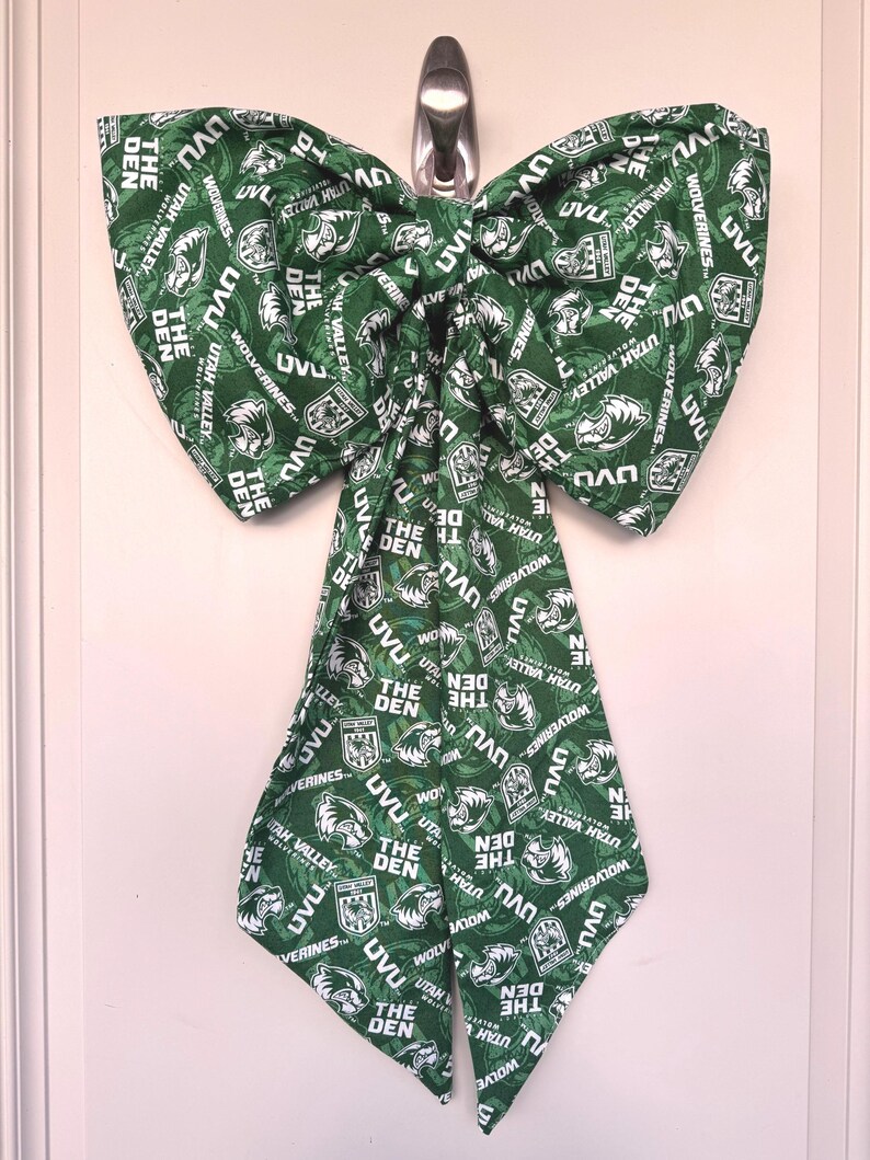 May include: A large green bow with a repeating pattern of white text and logos, including "UVU," "Utah Valley," and "The Den." The bow is hanging on a silver hook against a white door.