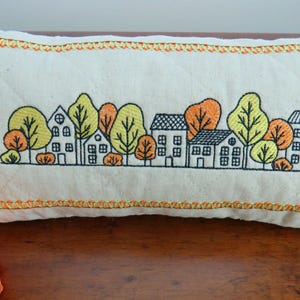 May include: Cream-colored rectangular pillow with an embroidered design of a village scene. The design features houses and trees in yellow, orange, and black. The pillow has a quilted pattern and is trimmed with orange stitching.
