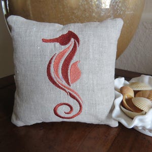 May include: A beige linen pillow with an embroidered red and orange seahorse design.