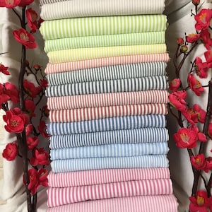 Op de afbeelding: Een stapel gestreepte stof in verschillende kleuren, waaronder bruin, wit, groen, geel, roze, rood, blauw en lichtblauw. De stof is netjes gevouwen en in een verticale stapel gerangschikt.