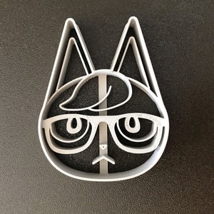 Puede incluir: Un cortador de galletas de pl&aacute;stico blanco con forma de gato con gafas. El gato tiene una cara redonda, ojos grandes y una nariz peque&ntilde;a. El gato lleva gafas con una montura negra gruesa.