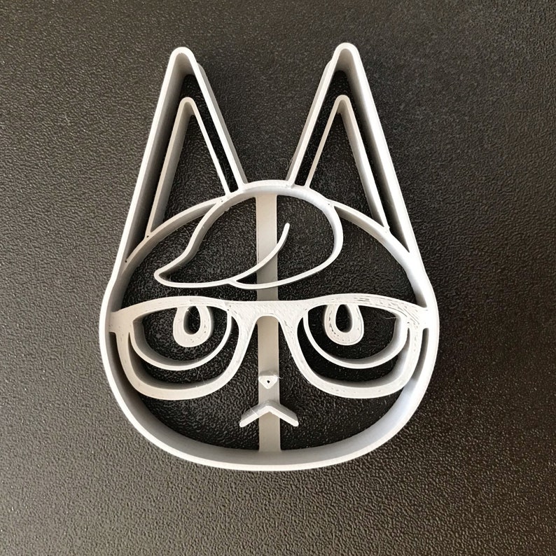 Puede incluir: Un cortador de galletas de pl&aacute;stico blanco con forma de gato con gafas. El gato tiene una cara redonda, ojos grandes y una nariz peque&ntilde;a. El gato lleva gafas con una montura negra gruesa.