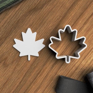 May include: Two white plastic maple leaf cookie cutters on a wood surface. One cutter is solid and the other is an outline.