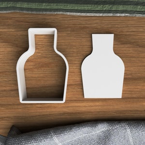 Puede incluir: Dos cortadores de galletas de plástico blanco con forma de botellas. Los cortadores están sobre una superficie de madera.