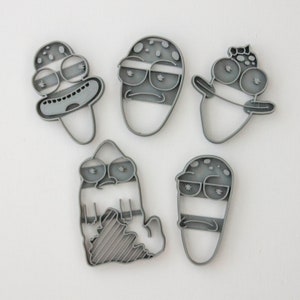 以下が含まれることがあります： 漫画のキャラクターの形をしたグレーのプラスチック製のクッキーカッター5個セット。キャラクターは大きな目、口、鼻をしています。