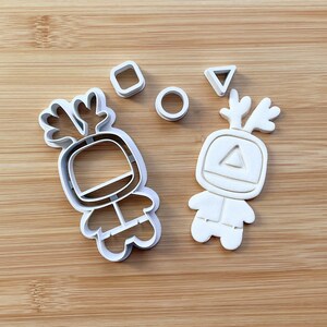 May include: Two white plastic cookie cutters shaped like cartoon characters with antlers. The characters are wearing spacesuits and have geometric shapes on their chests. There are also three smaller cutters shaped like a circle, square, and triangle.
