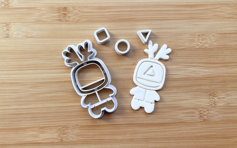May include: Two white plastic cookie cutters shaped like cartoon characters with antlers. The characters are wearing spacesuits and have geometric shapes on their chests. There are also three smaller cutters shaped like a circle, square, and triangle.