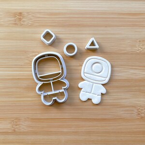 May include: A set of five white plastic cookie cutters. The cutters are shaped like a circle, a triangle, a square, and two astronauts. The astronauts are wearing spacesuits and have round helmets.