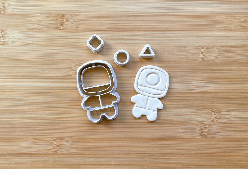 May include: A set of five white plastic cookie cutters. The cutters are shaped like a circle, a triangle, a square, and two astronauts. The astronauts are wearing spacesuits and have round helmets.