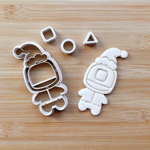 May include: Two white plastic cookie cutters shaped like astronauts wearing Santa hats. The cutters are on a wooden surface with a square, circle, and triangle cutter.