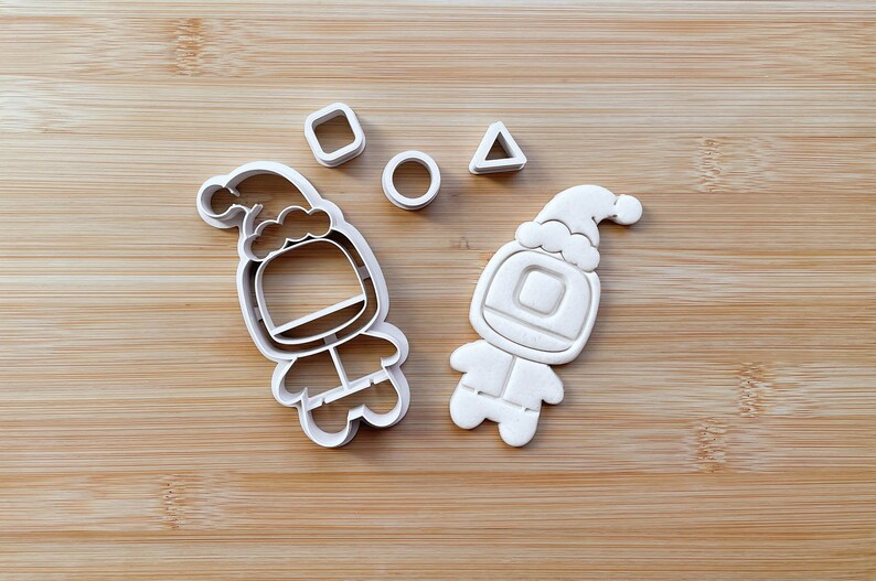 May include: Two white plastic cookie cutters shaped like astronauts wearing Santa hats. The cutters are on a wooden surface with a square, circle, and triangle cutter.