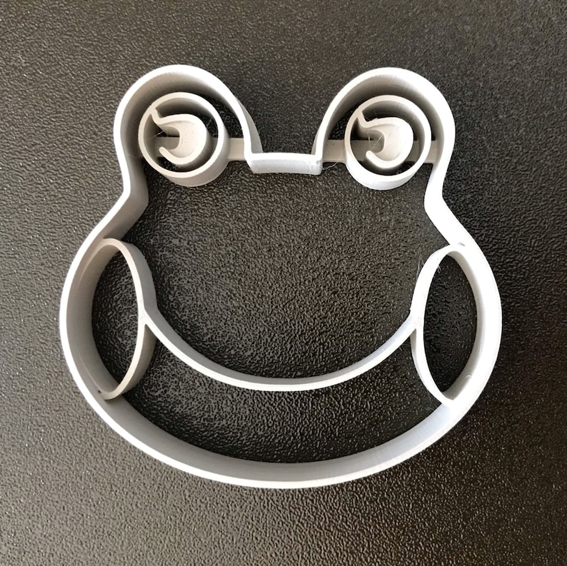 Puede incluir: Un cortador de galletas de pl&aacute;stico blanco con forma de rana con ojos grandes.