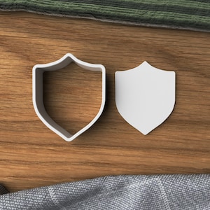 May include: Two white plastic cookie cutters shaped like shields. The cutters are on a wooden surface.