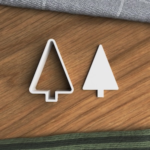 May include: Two white, Christmas tree-shaped cookie cutters on a wooden surface. One cutter is a solid shape, and the other is a frame. The cutters are designed for baking and crafting festive treats.