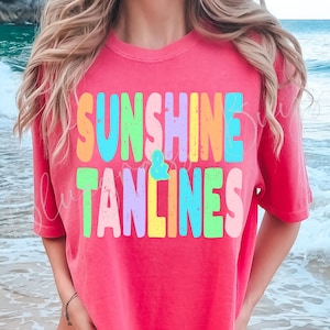 May include: A coral-colored t-shirt with the words "SUNSHINE & TANLINES" in a colorful, block letter design. The text is in shades of orange, blue, green, purple, and yellow. The shirt is a casual style.