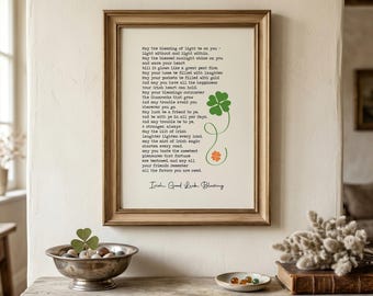 May include: A framed print featuring the Irish Good Luck Blessing text in black typewriter font. The design includes a green four-leaf clover graphic with a swirling stem ending in a small orange clover. The frame is a light brown wood finish.