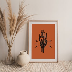 May include: A printable wall art design featuring a black skeleton hand making the rock and roll hand gesture with the text "Let's Get Spooky" in black on an orange background.