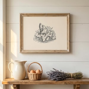 May include: A framed black and white print of a hen with chicks, displayed above a wooden shelf. The shelf holds a white pitcher, a wicker basket with eggs, and a bundle of lavender. The print has a light brown wooden frame.