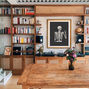 May include: A framed wall art print featuring a white skeleton illustration on a dark gray background. The skeleton is depicted from the chest up, making a rock and roll hand gesture with its left hand. The artwork is displayed in a gold frame.