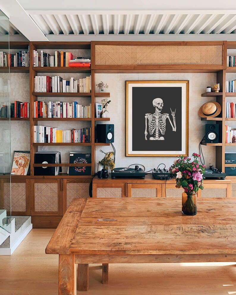 May include: A framed wall art print featuring a white skeleton illustration on a dark gray background. The skeleton is depicted from the chest up, making a rock and roll hand gesture with its left hand. The artwork is displayed in a gold frame.