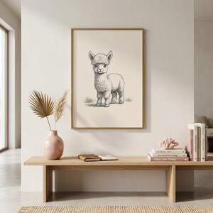 Vintage engraving-style baby alpaca wall art print featuring a detailed pencil sketch of an adorable fluffy alpaca cria standing and facing forward with a happy expression on a warm cream background, displayed in a gold frame in a bright minimalist boho living room with pampas grass and pink ceramic vase. Fine black linework captures the alpaca's fluffy coat and endearing expression beautifully. Perfect as alpaca wall art, boho home decor, or nursery animal art. Printable digital download