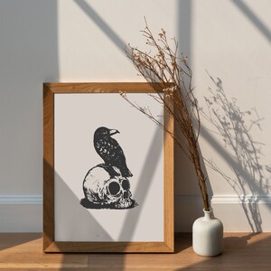 May include: A black and white illustration of a raven perched on a skull. The illustration is framed in a light brown wood frame and is hanging on a white wall. A dried flower arrangement is in a white vase on a wooden surface.