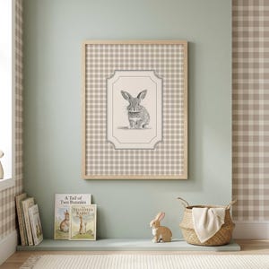 Könnte beinhalten: Ein gerahmter Druck mit einer Bleistiftskizze eines Kaninchens, vor einem beige-weiß karierten Hintergrund. Der Holzrahmen umgibt das Kunstwerk. Der Raum umfasst ein Regal mit Büchern, eine Kaninchenfigur und einen Weidenkorb.