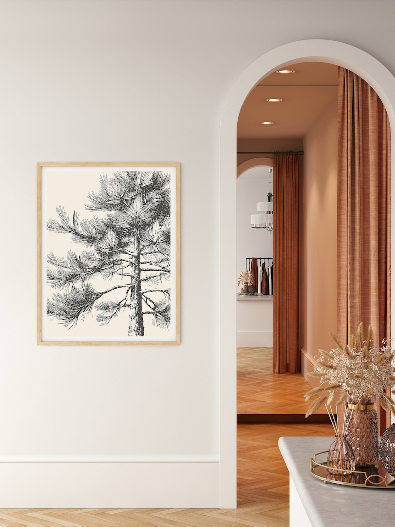 May include: A black and white line drawing of a pine tree in a light beige frame against a white wall. The frame is hanging on a white wall with a white baseboard. The wall is next to an arched doorway with a brown wall and a brown curtain.