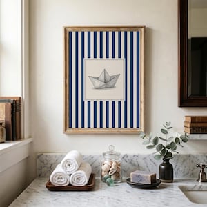 Op de afbeelding: Een ingelijst kunstwerk met een illustratie van een papieren boot. De achtergrond is blauw en wit gestreept, de lijst is van hout. De afbeelding toont een badkamer met opgerolde witte handdoeken en decoratieve elementen.