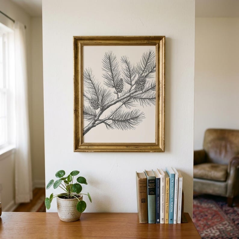 Vintage engraving-style pine branch wall art print featuring a detailed pencil sketch of a pine branch with three pine cones on a warm cream background, displayed in a gold ornate frame in a bright neutral living room. The fine black linework captures every pine needle and cone texture beautifully. Perfect as botanical wall art, farmhouse nature print, or cottagecore home decor for a living room, bedroom, or cabin. Printable digital download available in multiple sizes for instant home printing.