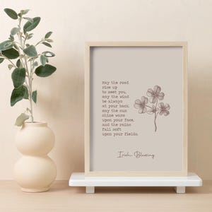 May include: A framed print with an Irish Blessing quote and a four-leaf clover illustration. The print is in a light beige frame and rests on a white shelf. A beige vase with greenery is to the left.