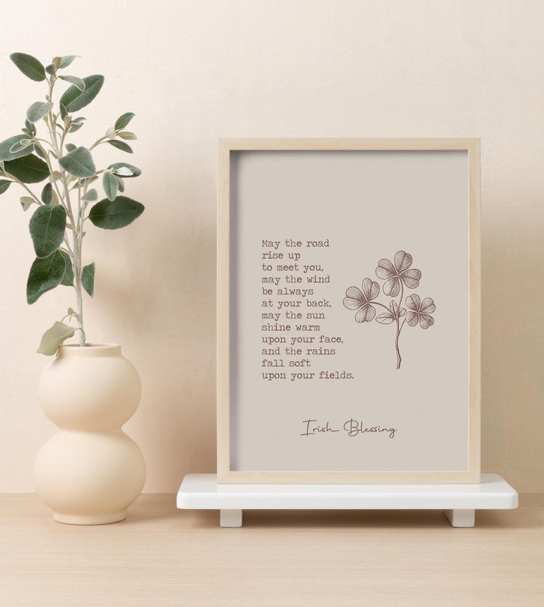 May include: A framed print with an Irish Blessing quote and a four-leaf clover illustration. The print is in a light beige frame and rests on a white shelf. A beige vase with greenery is to the left.