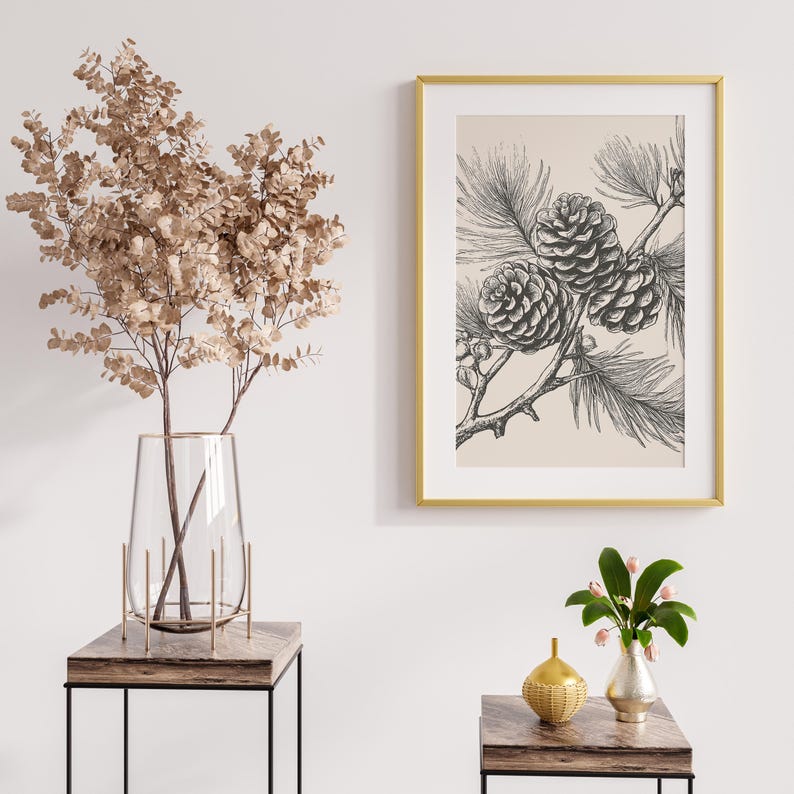 May include: A framed botanical print with a black and white illustration of pine cones and needles. The artwork is set against a cream background and framed in a gold-coloured frame. The image is displayed on a white wall.