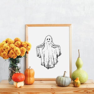 May include: A framed black and white illustration of a ghost with a smiling face. The frame is on a wooden table with a vase of yellow mums, gourds, and a green gourd.