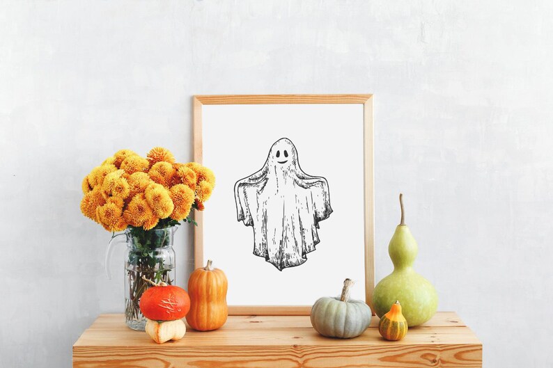 May include: A framed black and white illustration of a ghost with a smiling face. The frame is on a wooden table with a vase of yellow mums, gourds, and a green gourd.