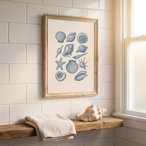 Puede incluir: Un estampado enmarcado con ilustraciones azules detalladas de conchas marinas sobre un fondo crema. La obra de arte está enmarcada en un marco de madera clara, evocando un tema costero. El estampado está montado en una pared de azulejos blancos.
