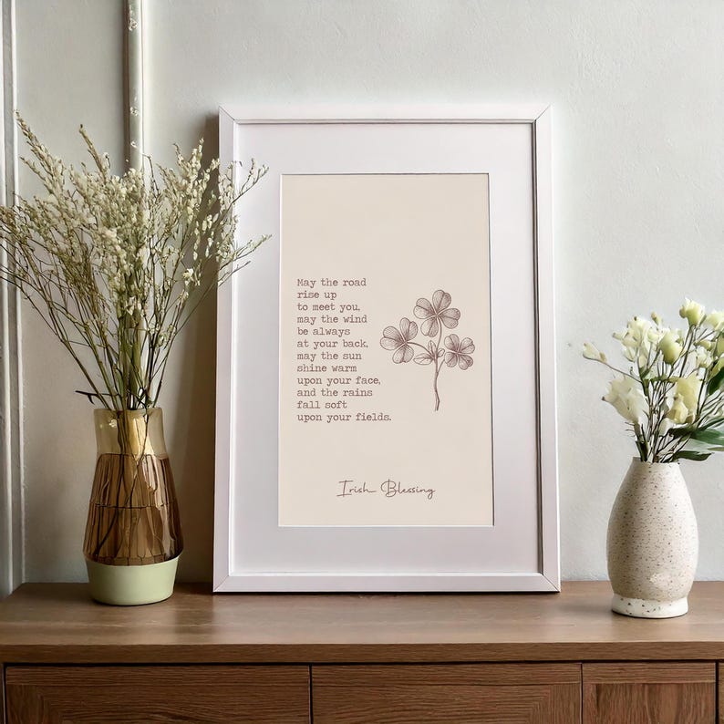 May include: A framed print with an Irish blessing in a white frame. The print has a beige background with a floral illustration and text. Two vases with flowers are on a wooden surface.