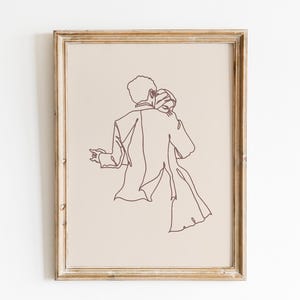 May include: A framed wall art print featuring a dark brown continuous line drawing of two figures embracing on a light beige background. The artwork is enclosed in a light-colored wooden frame with a visible grain, showcasing a couple in a dance.