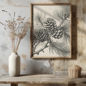 May include: A framed monochrome botanical print showing three detailed pine cones on a branch. The artwork is in a light wood frame, creating a rustic aesthetic. The print is displayed on a wooden surface.