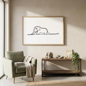 Puede incluir: Un cuadro con un dibujo lineal minimalista de un elefante negro sobre fondo blanco. La obra de arte está enmarcada en un marco de madera clara. El cuadro cuelga sobre un sillón verde y una mesa auxiliar de madera.