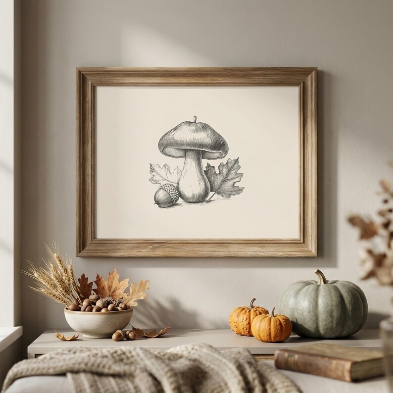 Vintage engraving style illustration of a mushroom with a rounded cap, flanked by oak leaves and an acorn, in a light wood frame on a warm beige wall above a console table with a bowl of acorns and dried leaves, wheat stems, and mini pumpkins. Printable digital download available in multiple sizes.