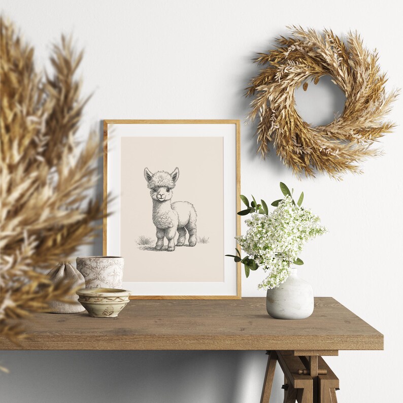 May include: A framed wall art print featuring a black and white sketch of a baby alpaca on a light beige background. The print is displayed in a light wood frame, positioned on a wooden surface next to a white vase with white flowers.