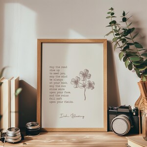 May include: A framed print with an Irish Blessing quote and a botanical illustration of clovers. The print is in a light wood frame and sits on a wooden surface. The text reads "May the road rise up to meet you...Irish Blessing."
