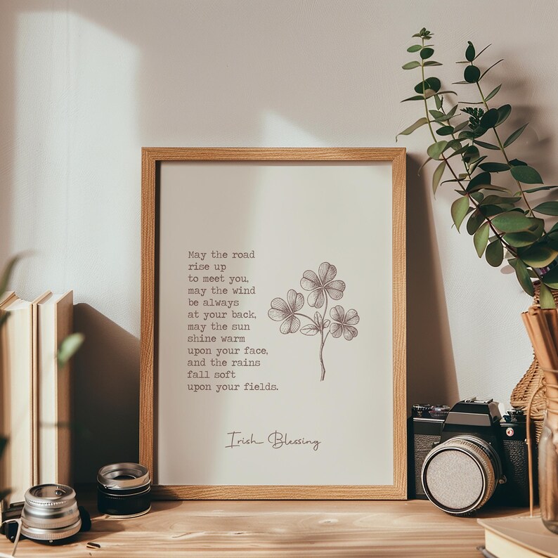 May include: A framed print with an Irish Blessing quote and a botanical illustration of clovers. The print is in a light wood frame and sits on a wooden surface. The text reads "May the road rise up to meet you...Irish Blessing."