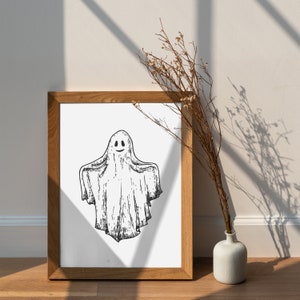 May include: A framed print of a black and white illustration of a smiling ghost. The ghost is drawn in a simple, cartoon style and is floating in the air. The print is in a wooden frame and is hanging on a white wall.