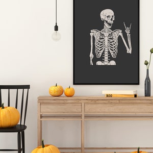 May include: A framed wall art print featuring a light-colored human skeleton illustration on a dark gray background. The skeleton is depicted from the waist up, with its left hand raised in a rock-and-roll hand gesture, set against a solid black frame.