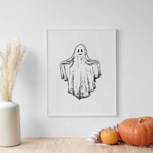 May include: A black and white illustration of a ghost with a smiling face in a white frame. The frame is on a white wall with a vase of dried pampas grass and two pumpkins on a wooden table.